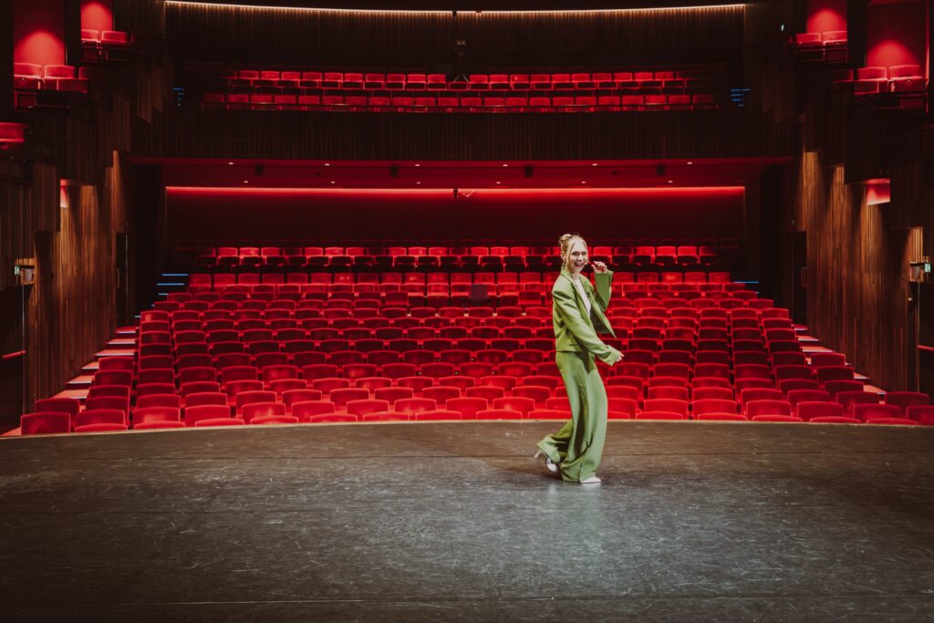 Manouk Schipper loopt sprekend over een theaterpodium in groen pak